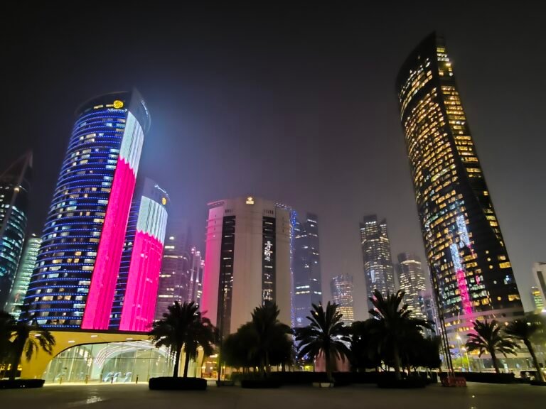 Nighttime Doha with high modern buildings, some covered in a projection of the magenta-and-white national flag.